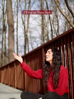 A woman with long wavy hair wearing a red sweater sits on a wooden platform with a wooden fence behind her and trees in the background. She has her right arm outstretched towards the sky.