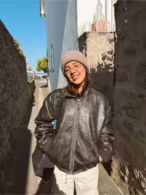 A woman is standing in a narrow alleyway. She is wearing a beige beanie, glasses, and a black leather jacket. The alley is made of stone walls, and there is a blue sky visible above. The image has a warm tone.