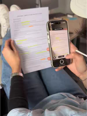 A person sits with their legs crossed, holding a document in one hand and a smartphone in the other. The document has highlighted text, and the phone's screen displays an app interface. The person is wearing jeans, white shoes, and a black sleeve.