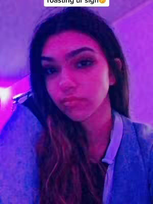 A selfie showing a young woman from the chest up. She has long, dark hair with some lighter streaks towards the ends, and her face is well lit, with a slightly posed expression. The background is filled with a vibrant, purplish-pink hue, with some lighter, brighter areas that suggest a light source.