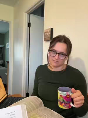 A woman with glasses holds a colorful mug, looking at the camera. She is sitting at a table with a laptop and an open book. The setting appears to be at home, in well-lit room.