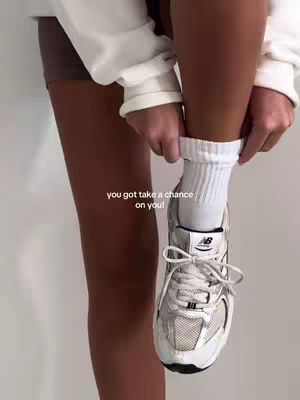 A person is putting on a white sock and tying a white and gray sneaker, the feet are in the frame and the video is a medium shot, on the bottom of the image appears the text 'you got take a chance on you!'