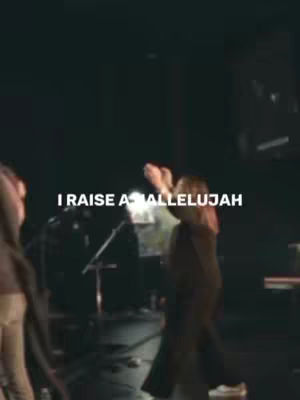 The image shows a performance, likely a concert or church service, with individuals on stage. The lighting is dim, potentially with spotlights, and the main text overlay states: 'I RAISE A HALLELUJAH'.