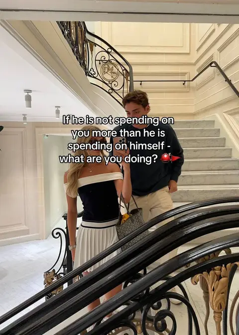 A well-dressed couple stands on a grand staircase; the woman is in a white skirt and black top, holding a phone, the man in neutral tones, with text overlayed.