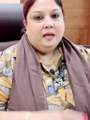 The image shows a woman in a close-up shot. She's wearing a patterned shirt and a scarf. She has dark hair and is looking at the camera with her mouth open. The background is out of focus.