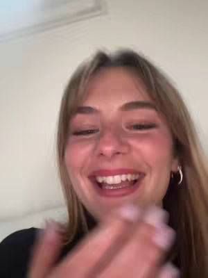 A selfie-style shot featuring a young woman with long blonde hair and a bright expression. She's facing the camera, with her eyes closed in a moment of laughter, and her hand is raised as if clapping. The background is a simple, light-colored wall or ceiling, with no other objects or details.