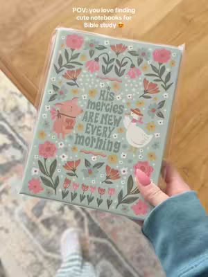 A hand holds a notebook with an illustrated cover in front of a soft patterned rug. The notebook is light blue-green with a pink and floral design. In the center, the notebook reads, "His mercies are new every morning." The hand holding the notebook has long, pink-painted nails. A portion of a light blue sleeve is visible.
