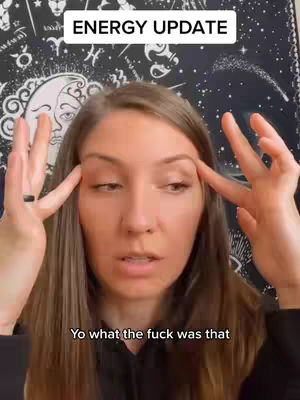 A woman with long brown hair is gazing up with a slightly confused expression. Her fingers touch her temples, as if to emphasize a thought, and a simple black ring adorns her left ring finger. Behind her, a patterned tapestry serves as the backdrop, its design obscured enough to leave the focus on the woman.