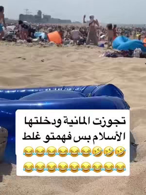 The image captures a crowded beach scene on a sunny day. The foreground features a large blue inflatable partially obscuring a sign with Arabic text and numerous laughing emojis. The background is filled with people, beach umbrellas, and buildings along the coast, suggesting a lively and bustling atmosphere.
