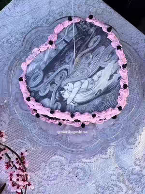 A heart-shaped cake sits on a patterned surface. The cake's design features a printed image depicting a scene from Junji Ito's Uzumaki, a spiral-themed horror manga. The cake is decorated with a pink frosting border, along with small dark-colored decorations. The background includes a patterned tablecloth and some of a floral display.