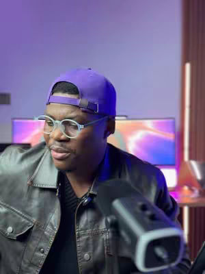 A man wearing a purple cap, glasses, and a black leather jacket is positioned in front of a desk with a microphone. The background has a gradient purple and orange color.