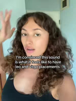 A woman with long, curly brown hair and minimal makeup is positioned centrally in the frame. She's wearing a black top and is mid-speech, her mouth open slightly. Her right hand is raised with fingers spread out, and the background is a light teal-green wall with a white door frame visible to the side.