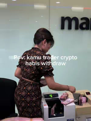 A woman in a floral dress is standing by a money-counting machine in a bank. She is partially turned away from the camera, appearing to count or organize bundles of cash. The setting looks like a financial institution with a large sign in the background.