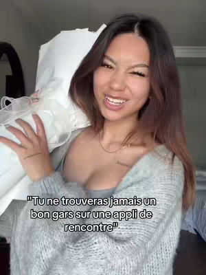 A young woman with long brown hair is smiling while holding a white item, possibly a pillow, against her chest. She's wearing a gray sweater. Text is overlaid on the image.