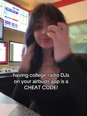 A woman with dark hair and bangs is the main focus, slightly out of focus, looking towards the camera while touching their face. The background shows a radio station control room with screens.