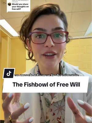 A woman with short, dark hair and glasses is speaking directly into the camera against a yellow wall. She is wearing a white lab coat over a patterned shirt. A text overlay reads "The Fishbowl of Free Will."