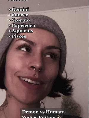 A woman with short, dark hair and a gray beanie looks toward the right with a slight smile. Text overlays include a list of zodiac signs and the phrase 'Demon vs Human: Zodiac Edition.'