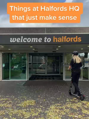 A person rides an electric scooter towards the entrance of a building with glass doors. The building has a sign that reads 'welcome to halfords' directly above the entrance. The top of the image has an orange rectangle with white text overlay.