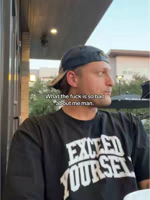 A man wearing a black t-shirt and a blue hat is captured from the chest up, gazing off to the side. The image is taken outdoors, with the man positioned against a backdrop of buildings and sky, suggesting an urban environment. Text overlayed on the image poses a question.