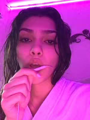 A close-up shot of a woman with dark hair, illuminated by a vibrant pink light from above. She holds the end of a white earphone in her mouth. The background is a wall with a decorative element.