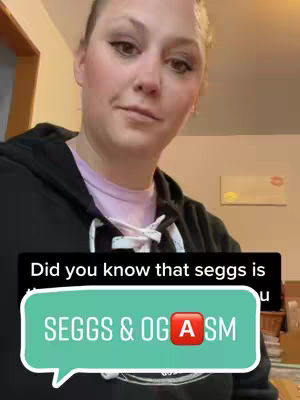 A woman is centered in the frame, looking slightly off-camera with a soft expression. She wears a black hoodie over a light pink shirt. Behind her, the background shows glimpses of an interior space, with neutral-colored walls and a glimpse of kitchen cabinets to the left. A speech bubble overlay reads "SEGGS & OG A SM," with a red emoji replacing a letter in the word "OGASM". Above the speech bubble a black text says "Did you know that seggs is..."