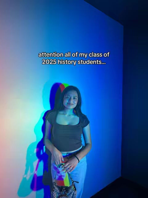 A young woman stands in front of a wall with a gradient of red, blue, and green light. She is wearing a brown top, light blue jeans, and is holding a purse. Text overlayed reads, "attention all of my class of 2025 history students..."
