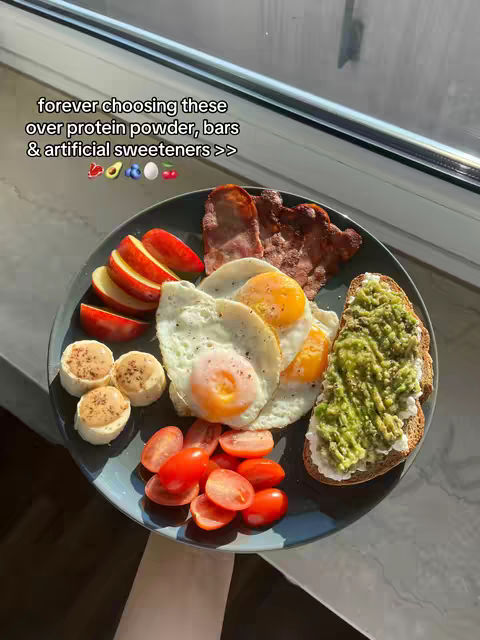 A plate of food is shown, including slices of apples, small biscuits, sliced tomatoes, fried eggs, bacon, and toast with a green spread. The plate is set in front of a bright window, and text is overlaid at the top.