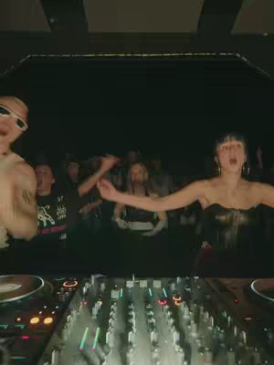 A chaotic, high-energy image from a rave or club setting. The focus is on a DJ at the front with a crowd of dancing people behind them. The lighting is dim and creates a vibrant and dynamic atmosphere.