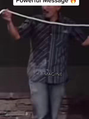 A man in a button-up shirt stands with arms outstretched, holding a white rope. The background is dark.