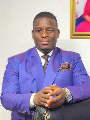 A man in a blue suit poses, hands clasped in front of him. He has a brown tie, and a patterned pocket square. A painting is in the background to his right.