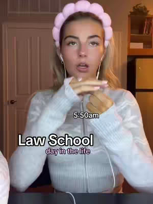 A young woman with blonde hair wearing a light blue sweater and a pink headband is the central focus against a warm-toned room backdrop. Text overlays include "Law School day in the life" in a bold font and "5:50am" indicating the time. The woman is looking at the camera. The background features a closed white door and a partial view of a wooden cabinet, suggesting an indoor setting.