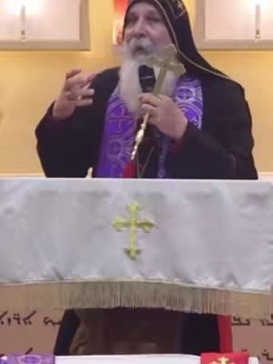 The cover image features a man, likely a religious figure, speaking at a podium. He wears religious attire and holds a cross. The background is a modest interior space, with a white tablecloth in front of him bearing the golden cross symbol. The image has warm lighting with a slightly blurred background