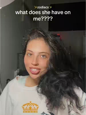 A woman with fair skin and long dark hair is the central figure in the shot, positioned from a waist-up perspective. Her mouth is slightly open in a partial smile, and she has visible upper teeth. She wears a white sweatshirt with a large gold crown design. The background shows a dimly lit room with a dark gray wall. A dark screen, possibly a TV, is in the background to the upper right.