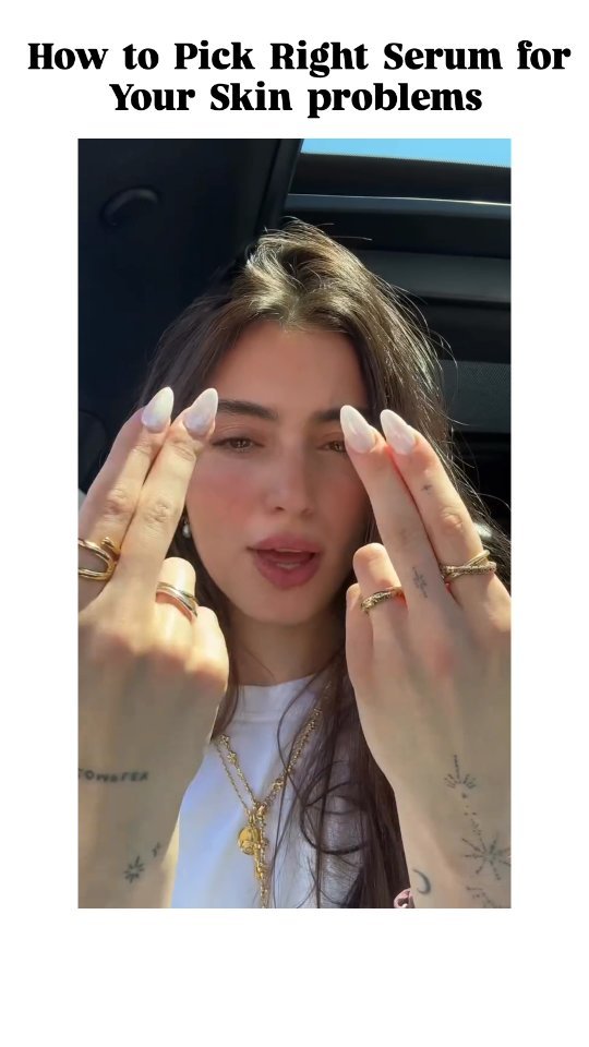 A woman sits in the front seat of a car with her hands in front of her face, both hands have the index and middle fingers up. She is wearing multiple rings on her fingers and a necklace. The top of the car is visible as is the outdoor background behind her.