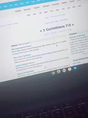 A screen of a website displaying a passage from 1 Corinthians 7:9. The site has a blue header with multiple tabs at the top. The main content is white with black text. A person's hands and the corner of a laptop keyboard are visible at the bottom.