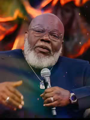 The image shows a Black man with glasses and a white beard holding a microphone. He is wearing a dark blue suit, and an ornate watch is on his wrist. The background is blurry with warm colors, suggesting a stage or studio setting.