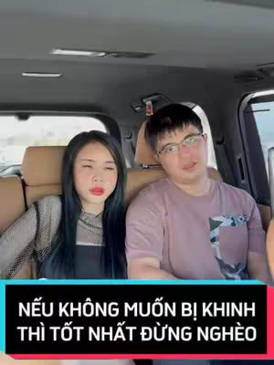 Two people, a woman and a man, are seated inside a car. The woman, on the left, is wearing a black top with her long, dark hair styled down. The man on the right wears glasses and a light-colored shirt. A text overlay at the bottom reads a Vietnamese phrase.