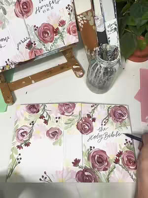 The image showcases a hands-on painting project in a studio setting. A partially opened the Holy Bible is spread across the bottom half, its cover painted with watercolor roses in various shades of pink and burgundy, accented with green leaves. The words 'the Holy Bible' are handwritten in elegant script on the cover. The artist is applying ink with a pen. To the left, another Bible sits on a small wooden easel, the cover similarly decorated. Above and to right, a glass jar holds paintbrushes. The background includes additional art supplies and a glimpse of a green plant, all on a white surface. The composition is well-lit and showcases the creative process of hand-painted Bible art.