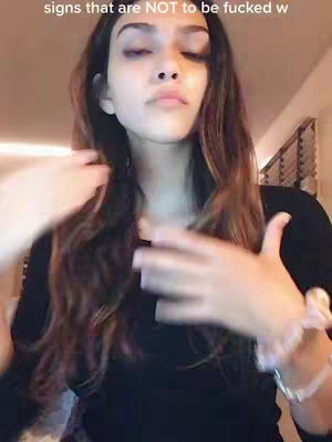A young woman with long, wavy brown hair is the central focus, her face slightly tilted upwards with a somewhat serious expression. She's wearing a black top and has a light-colored bracelet on her wrist. Her hands are raised near her face as though gesturing. The lighting is soft, with a slightly blurred background.