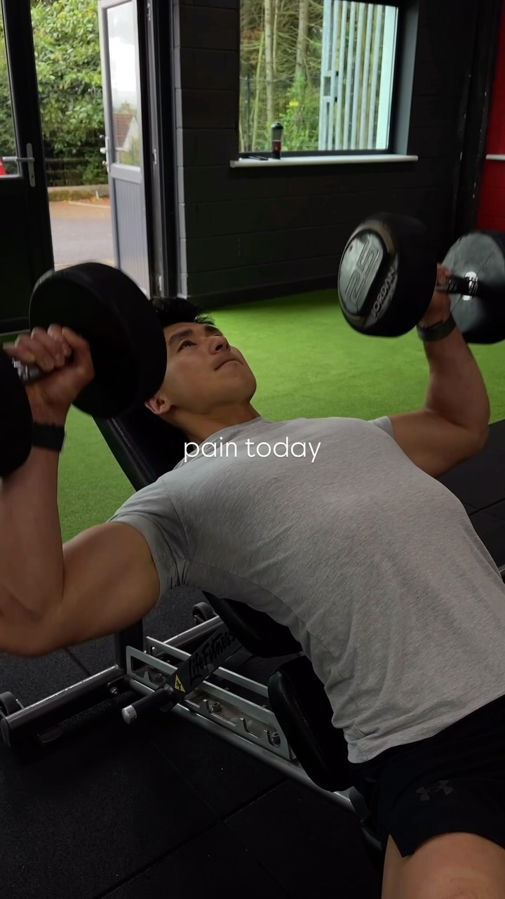 A man is lying down on a weight bench, lifting dumbbells overhead. He is wearing a grey t-shirt and black shorts. The image is taken from a low angle, looking up at the man. The text overlay says 'pain today'.