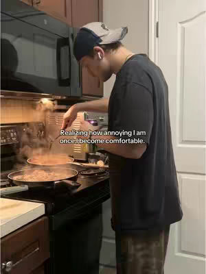 A man in a kitchen cooking food on a stove. Text overlayed on the image reads: 'Realizing how annoying I am once I become comfortable.' The scene is lit by an overhead light, creating a warm tone. The man is wearing a baseball cap and a dark t-shirt, with steam rising from the pans on the stove.