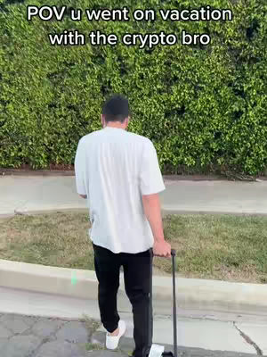 A man in a white t-shirt and black pants, walks away from the camera, holding a suitcase. The background is a wall of green bushes. Text overlay says 'POV u went on vacation with the crypto bro'.