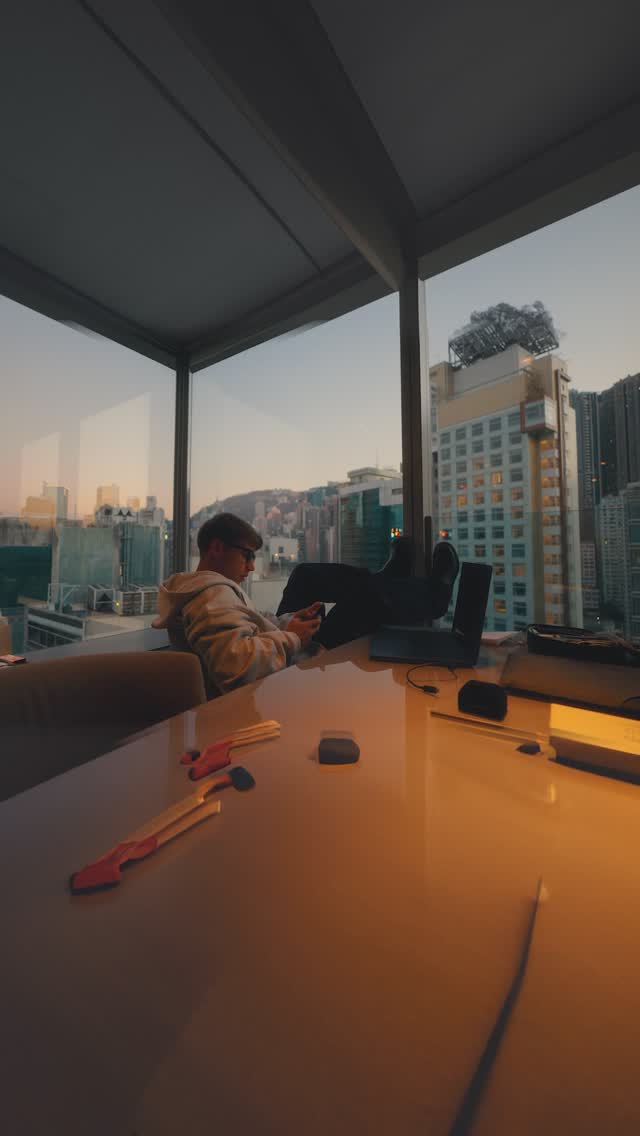 A person is seated at a table with their feet up, looking at a phone in a high-rise room overlooking a city. The composition is balanced, with a focus on the subject and the urban background, with an ambient warm lighting.