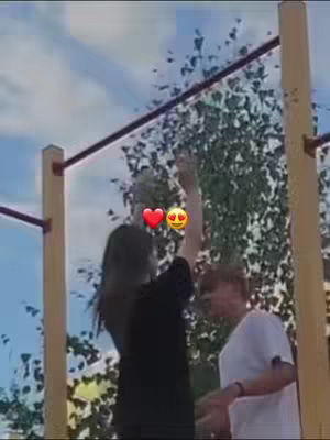 A sunny outdoor shot featuring a woman hanging from a horizontal bar, with a man standing nearby. Emojis are added near the woman's head. Green foliage and the sky create a natural backdrop.