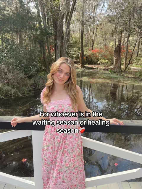 A young woman with long blonde hair stands outside, smiling. She's wearing a pink floral dress and holding her arms out to the sides. Behind her, there's a blurred background of trees and a body of water. Overlayed on the image is white text in a black rectangle.