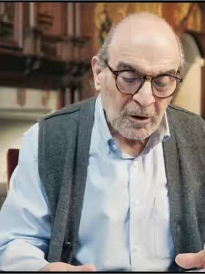 An older man with glasses in a light blue shirt and grey sweater vest is looking down with a slight open mouth. The background is blurred with various shades of brown.