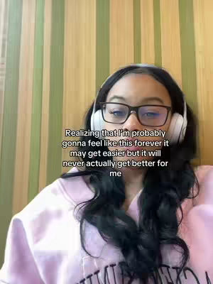 A selfie-style shot features a young woman with long, dark hair, wearing glasses and headphones around her neck. She's positioned against a background of vertical green and brown striped panels. Overlayed text at the bottom reads, 'Realizing that I'm probably gonna feel like this forever it may get easier but it will never actually get better for me.'