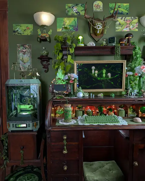 A cozy, whimsical desk setup with a rolltop desk and green accents. A large open desktop computer with a green glow occupies the left side. A detailed desk with various decorations: plant life in pots, printed artwork, and a green-lit keyboard. The color scheme is dominated by shades of green and brown, with a whimsical aesthetic. The background is a green painted wall, with additional artwork and ornaments. Plants and nature-themed aesthetics pervade the entire scene.