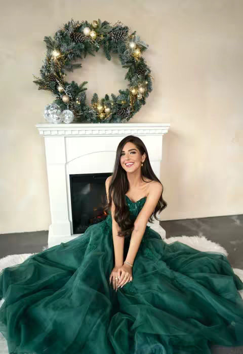 A woman in a formal green dress smiles in front of a decorated fireplace. A large wreath of greenery and fairy lights is above the fireplace. The woman has long dark hair. The dress has a voluminous skirt, and she is sitting on a fluffy white rug.