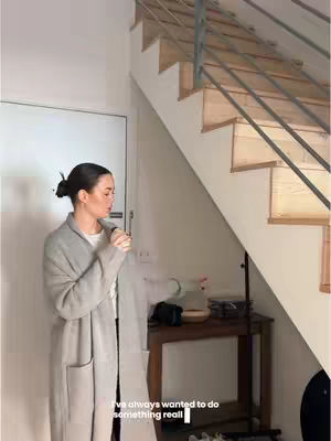 A woman in a grey coat stands next to a white door and wooden staircase.